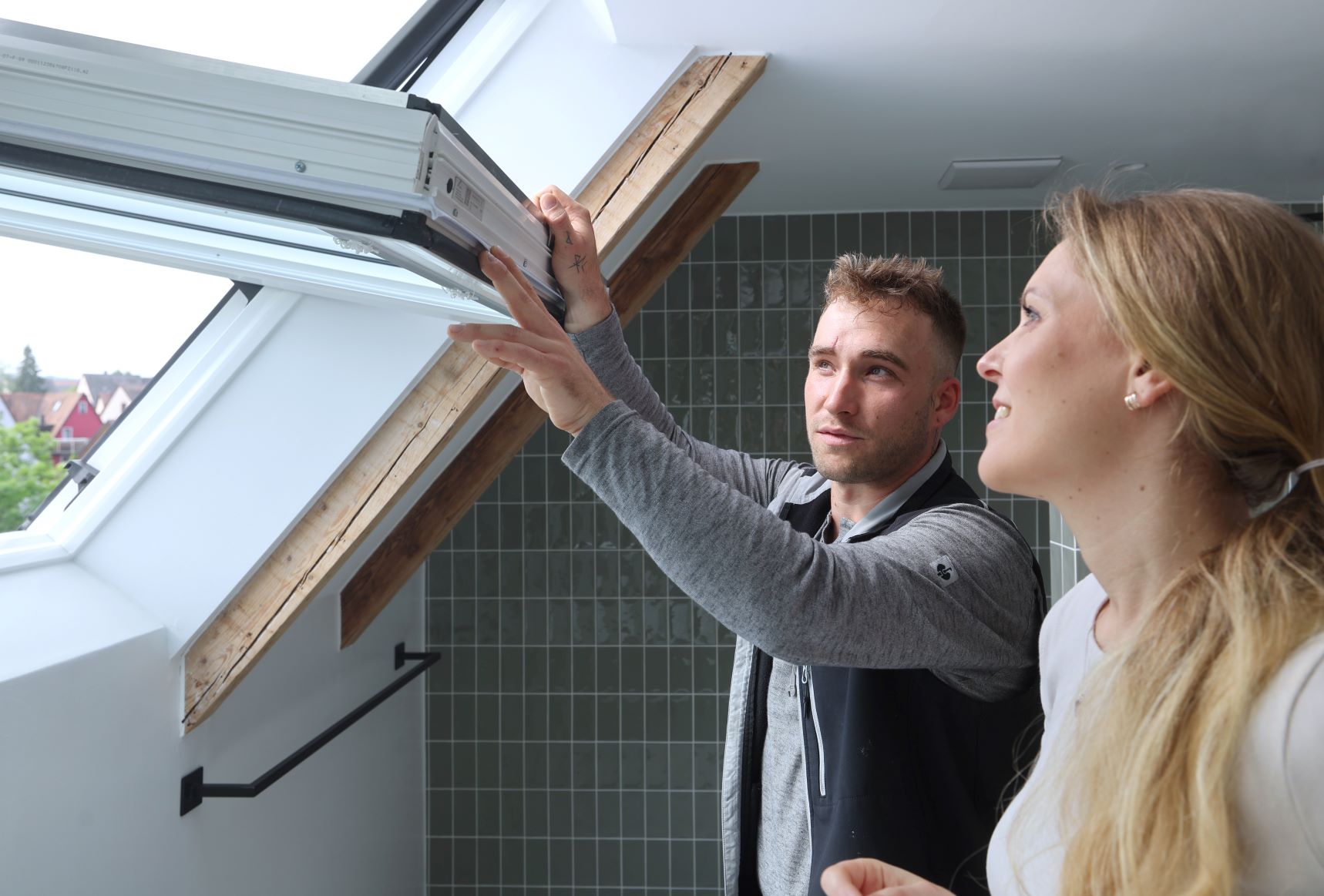 Ein Servicetechniker und eine Kundin stehen vor einem geöffneten Dachfenster. Der Servicetechniker überprüft das Fenster.