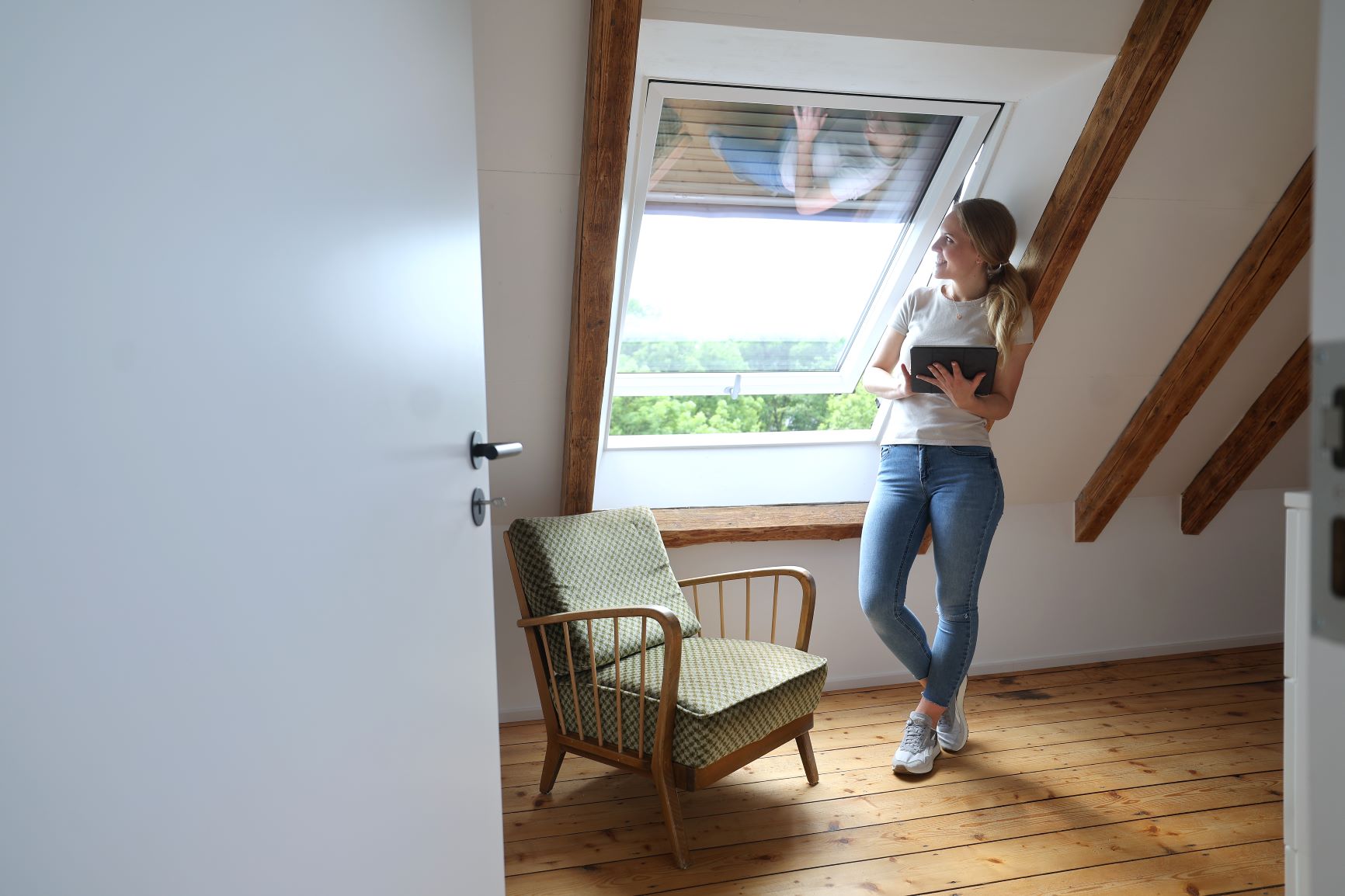 Kundin steht mit ihrem iPad in der Hand vor Ihrem automatisierten Dachfenster und blickt nach draußen.