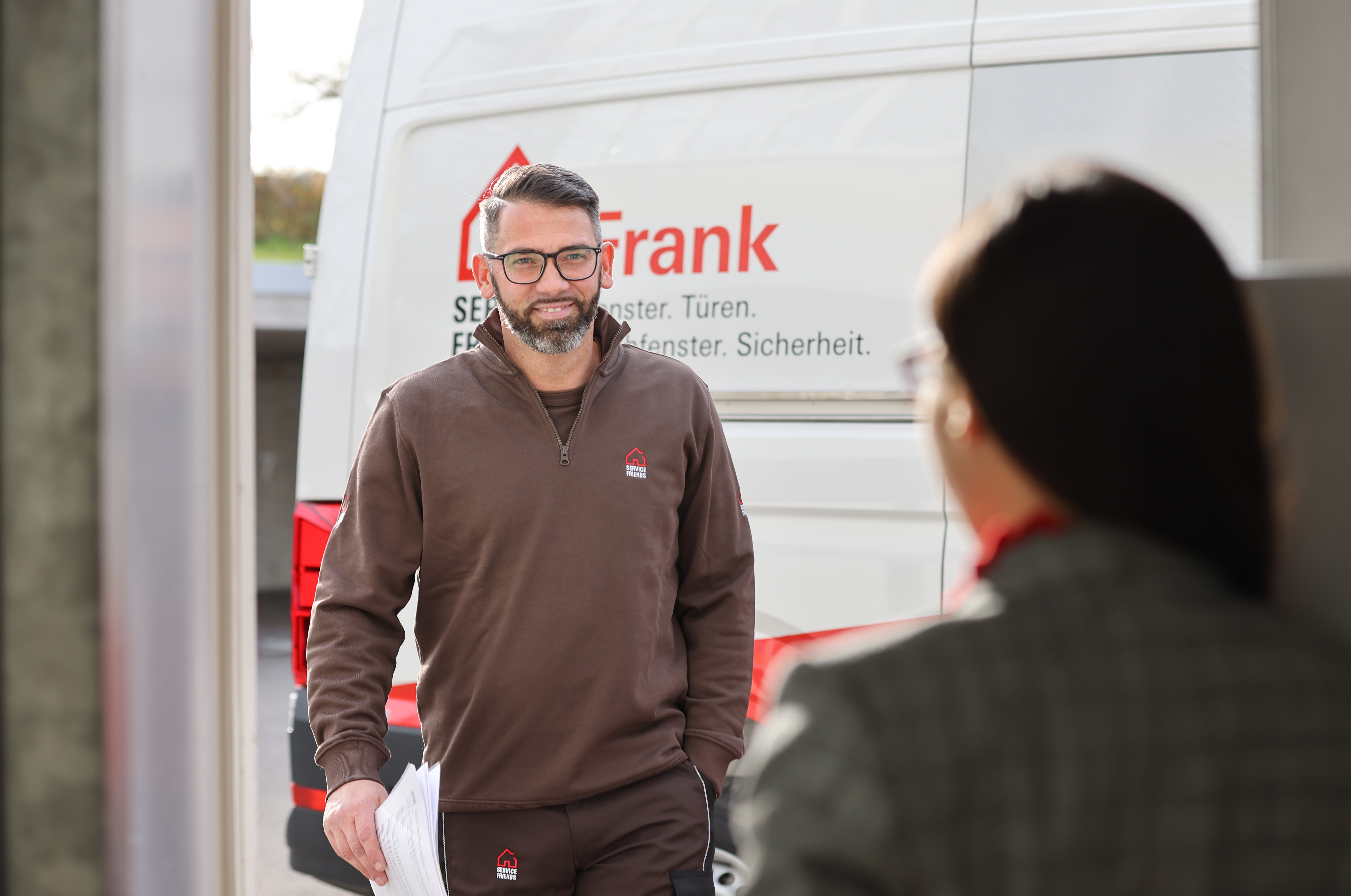 Ein Servicetechniker kommt gerade bei einer Kundin an, die mit dem Rücken zur Kamera steht. Im Hintergrund sieht man ein Firmenfahrzeug der Frank Sicherheits- und Fenstertechnik GmbH.