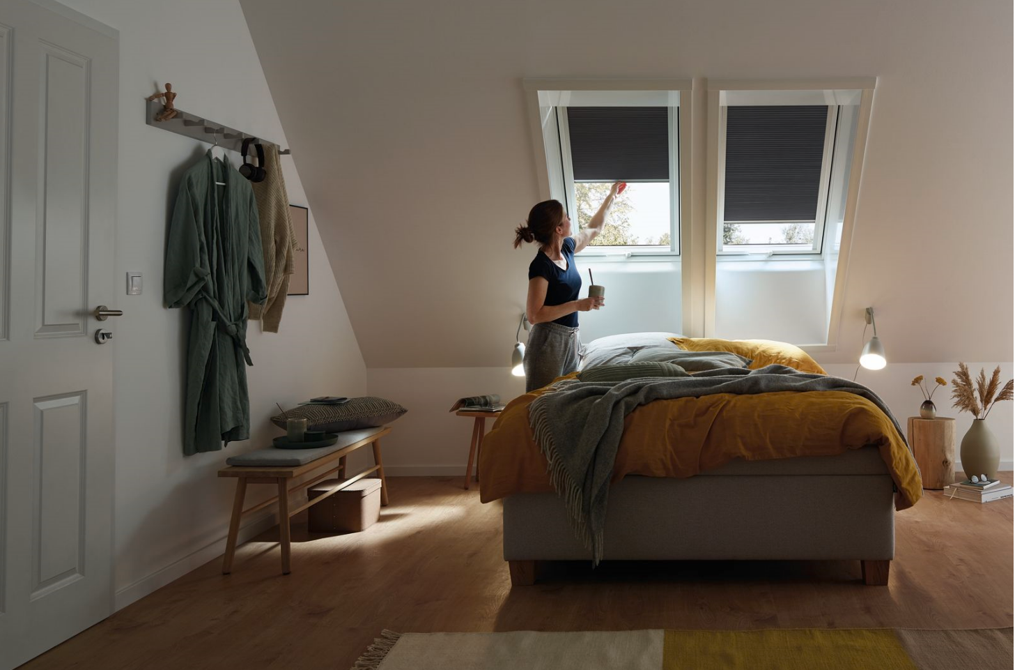 Eine Frau in Ihrem Schlafzimmer. Man sieht ein großes Bett unter einer Dachschräge mit zwei Dachfenstern. Die Dachfenster haben Verdunkelungsrollos. Die Frau steht vor einem der Fenster und schiebt den Rollo nach oben.