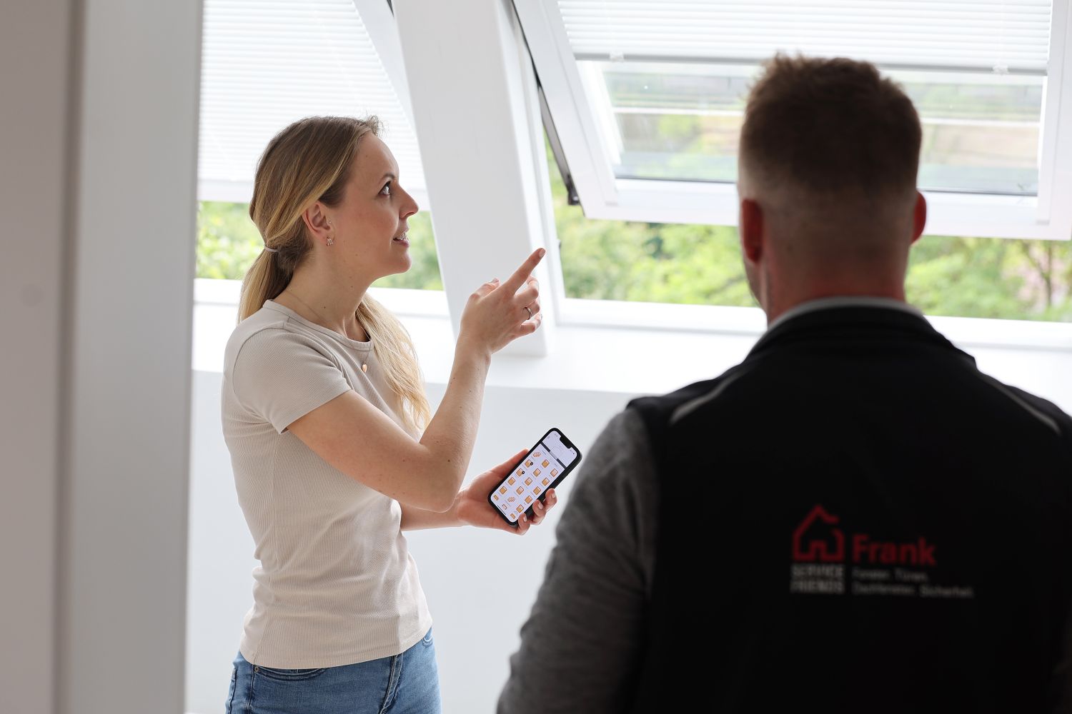Eine Kundin zeigt auf ein geöffnetes Dachfenster und hält dabei ein Smartphone in der Hand. Ein Serviceterchniker steht mit dem Rücken im Bild, während er ihr etwas erklärt.
