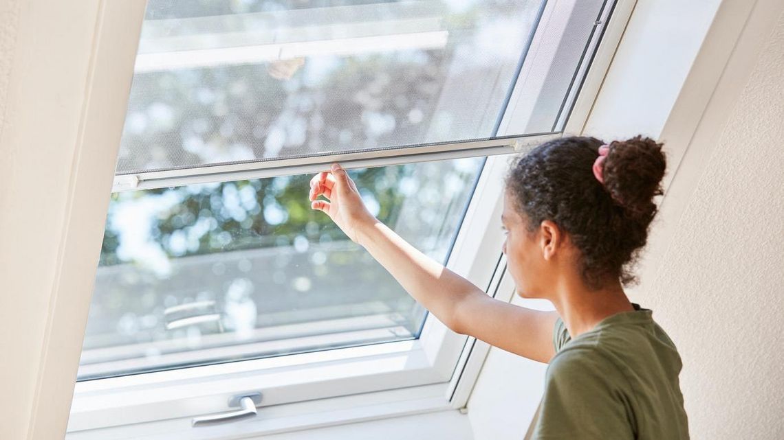 Eine Frau schließt ein Insektenschutzrollo, das vor einem Dachfenster angebracht ist.