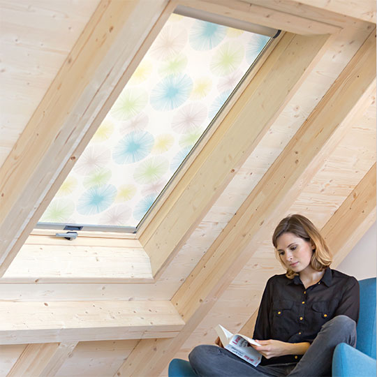 Eine Frau sitzt auf einem Sessel neben einem Dachfenster und liest ein Buch. Das Dachzimmer ist mit hellen Holzbalken verkleidet.