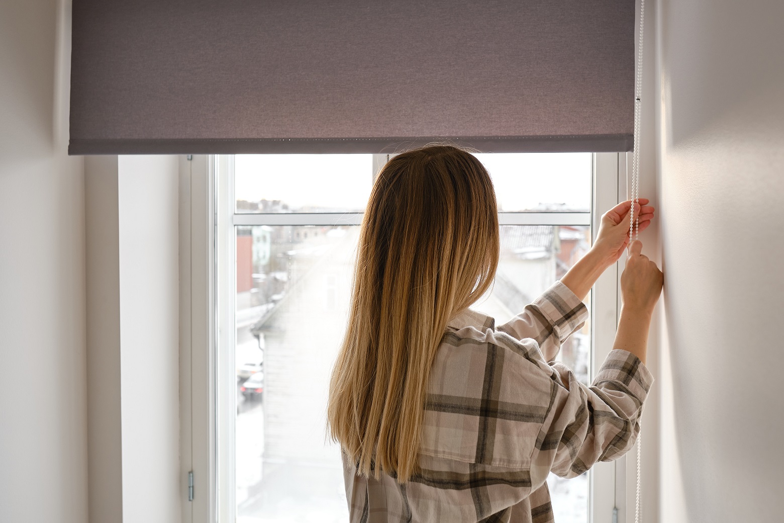 Eine Frau steht vor einem Fenster und zieht an einer Kordel, um ein Rollo herunterzulassen.